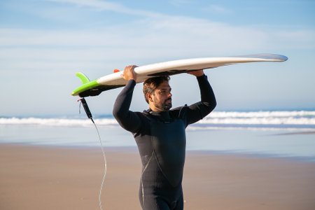 Male surfer in wetsuit walking on beach and carrying surfboard overhead. Medium shot. Surfing and...