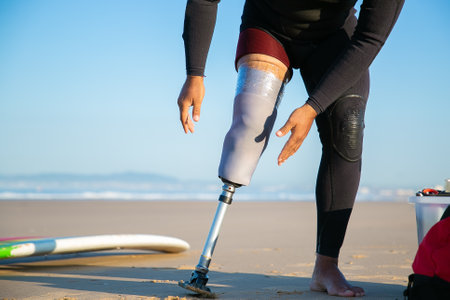 Male surfer wearing wetsuit for, adjusting artificial limb taped to leg. Cropped shot. Artificial limb and active lifestyle conceptの写真素材