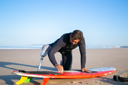 Sporty man in wetsuit wearing artificial limb, waxing surfboard on sand on ocean beach. Artificial limb and active lifestyle conceptの写真素材
