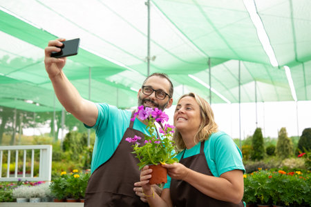 Bearded man and blonde woman doing selfie with petunia and smiling. Male gardener in eyeglasses holding phone and standing with colleague in greenhouse. Gardening and digital technology conceptの写真素材