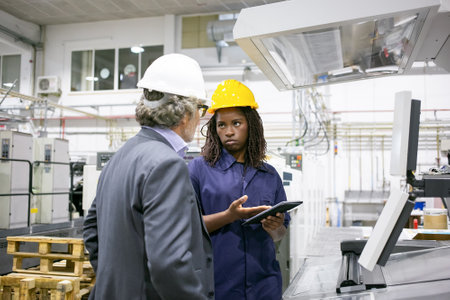 Serious black female factory worker talking to boss at machine on plant floor, pointing at tablet...
