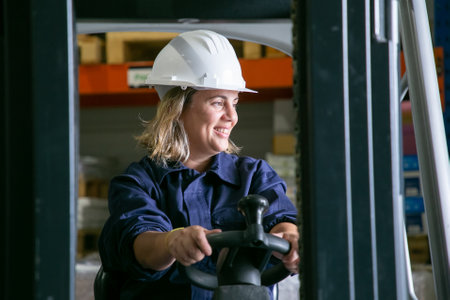 Happy Caucasian female logistic worker in hardhat driving forklift in warehouse, smiling, looking away. Side view. Female labor conceptの写真素材