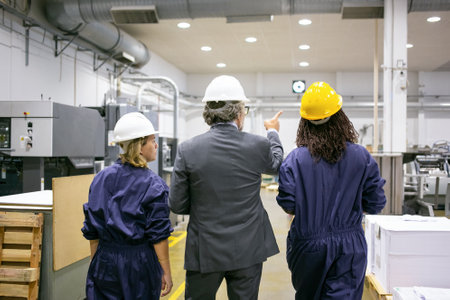 Male engineer and female factory employees in hardhats walking on plant floor and talking, man...
