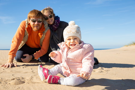 Cheerful couple watching cute baby girl wearing warm clothes, sitting on sand outdoors. Leisure time...