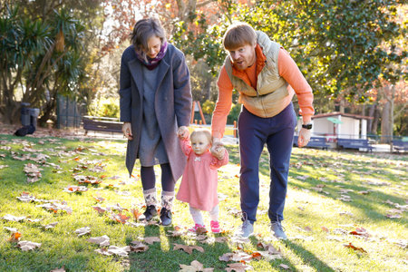 Happy parents walking with little girl in fall park and holding her hands. Lovely toddler in pink dress doing first steps on grass with help of bearded dad and blonde mum. Family and holiday conceptの写真素材