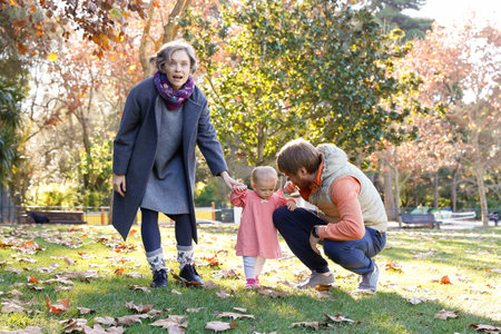 Cheerful family with little girl walking in autumn park on lawn. Blonde mum standing with open mouth and looking forward. Bearded dad squatting and talking with kid. Parenthood and holiday conceptの写真素材
