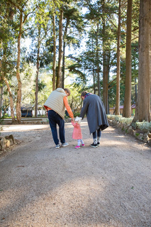 Family couple holding hands of baby girl and walking in autumn park. Kid turning around and looking at camera. Back view, full length. Family and childhood conceptの写真素材