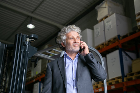 Pensive mature businessman standing near forklift in warehouse and speaking on cell phone. Shelves with goods in background. Business or logistics conceptの写真素材