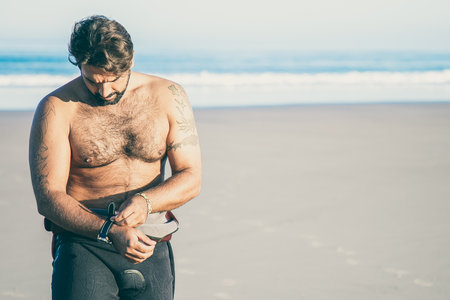 Sportsman putting on wetsuit for surfing on ocean beach and taking off watch. Copy space, front...