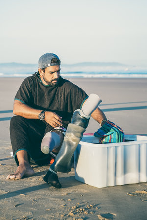 Focused male surfer sitting on beach and changing below-knee prosthesis. Front view. Artificial limb...