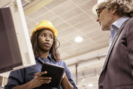 Serious black female factory employee talking to boss at her workplace on plant floor, using tablet...