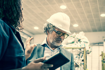 Mature male factory engineer talking to black female worker on plant floor at control board of machine. Copy space. Production process or machinery conceptの写真素材
