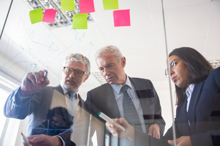 Focused grey haired male business leader drawing chart on glass board, explaining project details to colleagues, showing diagram. Teamwork or analysis conceptの写真素材