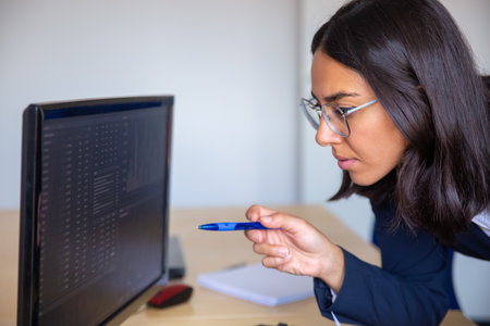 Focused female finance professional pointing pen at statistic report on monitor, analyzing stock...