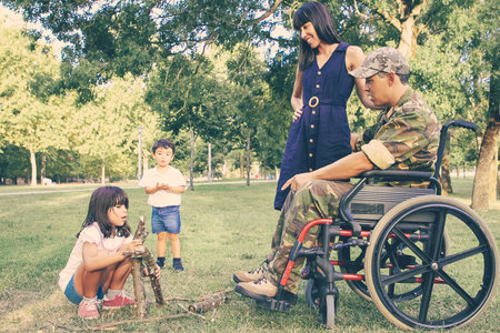 Cheerful mom and disabled military dad in wheelchair looking at kids arranging firewood for campfire outdoors. Disabled veteran or family outdoors conceptの写真素材