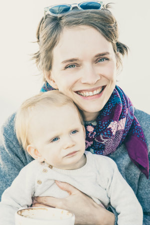 Cheerful mom holding sweet baby in arms while drinking coffee in coffee shop. Leisure time or coffee break conceptの写真素材