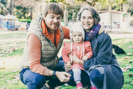Happy dad, mum and daughter posing for photo in autumn park and squatting on grass. Blonde beautiful...