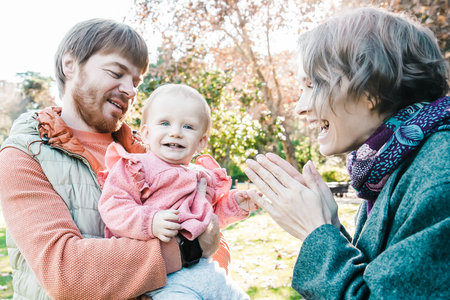 Happy mom and dad playing with cute baby girl in autumn park, holding kid who looking at camera and...