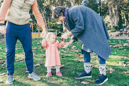 Happy parents with little girl walking in autumn park on lawn. Caucasian mom and dad holding cute...