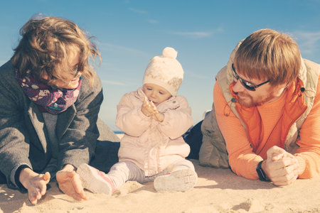 Relaxed couple of parents and cute baby daughter wearing warm clothes, sitting and lying on sand together outdoors. Leisure time and parenthood conceptの写真素材