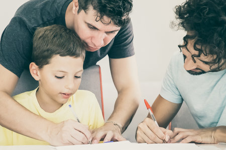 Gay parents helping focused boy with school home task, sitting at table and writing in papers together. Family and parenthood conceptの写真素材