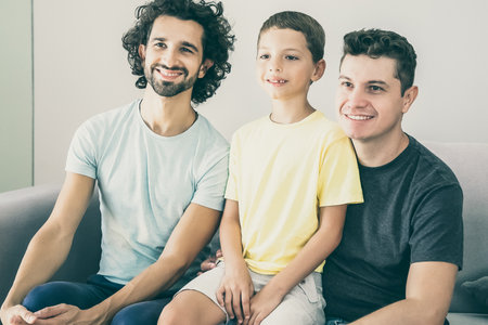 Happy gay fathers and child sitting on couch at home, smiling, and looking away. Front view. Family and parenthood conceptの写真素材