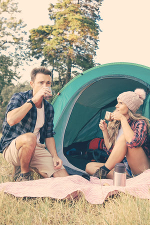 Young tourists sitting in tent and drinking tea from thermos. Caucasian travelers camping on nature and relaxing on lawn together. Backpacking tourism, adventure and summer vacation conceptの写真素材