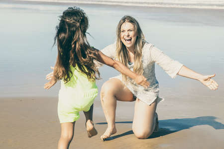 Happy excited mom opening arms for catching and hugging little daughter. Girl running to mother on beach at sea. Parenting and outdoor activities conceptの写真素材