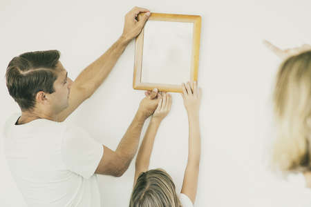 Father and daughter hanging up frame with copy space on wall in their new home. Blonde mother...