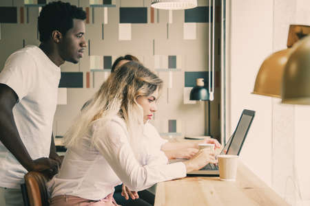 African American guy standing behind blonde woman and looking at screen. Caucasian lady sitting at table and working with laptop computer. Communication, digital technology and teamwork conceptの写真素材