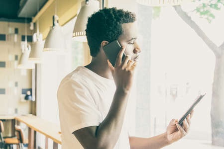 Serious African American guy talking on cell and looking at tablet screen. Young man using gadgets...