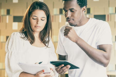Serious African American client looking at woman notebook and holding tablet. Pretty brunette female...