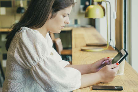 Serious pretty young woman in white shirt using tablet while sitting at desk in co-working space or coffee shop. Side view. Wireless technology conceptの写真素材