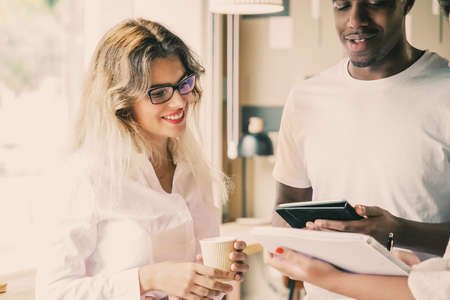 Beautiful young lady in glasses holding plastic cup, smiling and looking at notebook. Caucasian blonde woman standing near African American man with tablet. Communication and teamwork conceptの写真素材