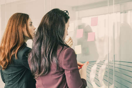 Concentrated businesswomen sticking notes on glass wall in conference room. Successful confident female employees working together and preparing for brainstorming. Business and management conceptの写真素材