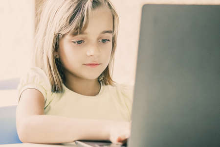 Caucasian pretty girl typing on laptop computer. Concentrated child sitting at table, browsing and studying online via electronic device. Childhood, communication and digital education conceptの写真素材