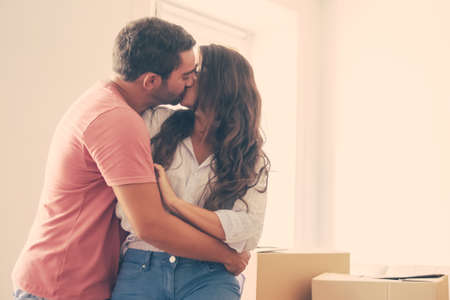 Young family couple moving into new house, standing near heap of carton boxes and kissing. Medium shot, copy space. New home and love conceptの写真素材