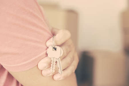 Man holding key from his new apartment, standing with arms folded and watching his girlfriend carrying carton boxes. Cropped shot, closeup. Property buying or mortgage conceptの写真素材