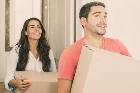 Joyful young Latin couple coming in their new apartment with carton boxes. Medium shot. Moving or...