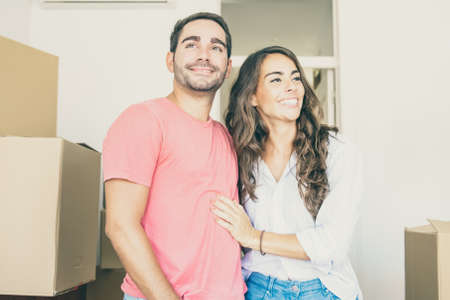 Cheerful couple looking over their new apartment, walking among carton boxes and hugging. Medium...