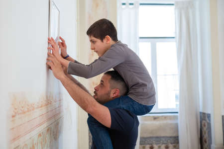 Concentrated son and bearded father hanging picture on wall. Caucasian boy sitting on mans neck, looking at picture, decorating apartment during renovation. Side view shot. Family concept.の写真素材