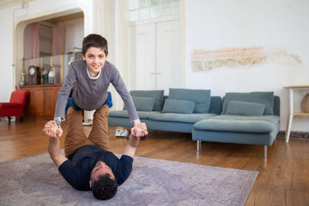 Cheerful single dad lying on floor, lifting his son. Caucasian boy imagining he flying as airplane. Dad playing with kid at home. Fatherhood, family, having fun togetherの写真素材
