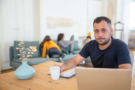Concentrated father sitting at table, looking at camera. Bearded dad working at home, using laptop while his children playing. Fatherhood, work at home conceptの写真素材