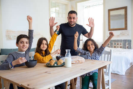 Happy dad with kids sitting in kitchen, rising their hands up. Kids enjoying their delicious breakfast meal. Breakfast, fatherhood, family conceptの写真素材