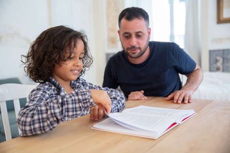 Curly-haired boy sitting with father at table and studying. Cheerful son turning page of textbook on, looking at it. Bearded father helping kid with homework. Reading book together, family conceptの写真素材