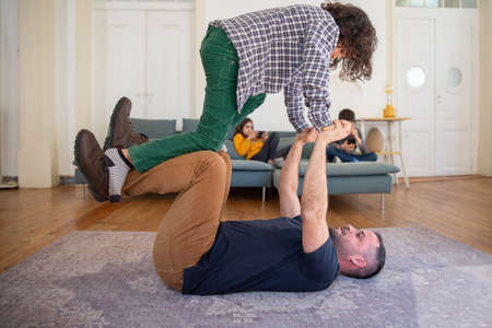 Happy dad lying on floor and lifting his son. Curly-haired boy balancing on daddys hands and happy...