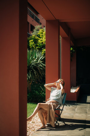 Pretty mid adult woman resting on veranda outdoors. Lady wearing sunglasses sitting barefoot on chair and sunbathing. Resting outdoors conceptの写真素材