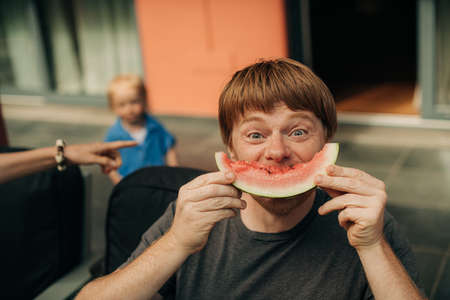 Portrait of joyful mid adult man eating watermelon. Father resting with his family at home and...
