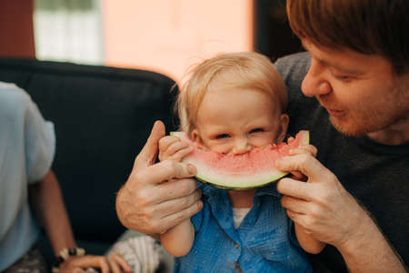 Frowning little toddler eating slice of watermelon. Mid adult father feeding his daughter with piece...