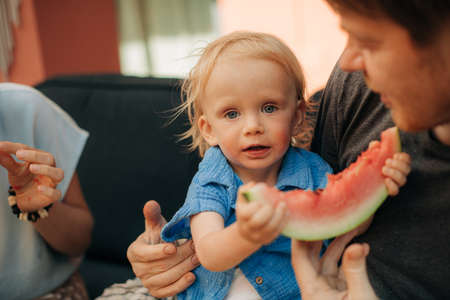 Close-up of cute little girl holding watermelon. Adorable toddler sitting on her father lap near her mother and looking at camera. Childcare and childhood conceptの写真素材